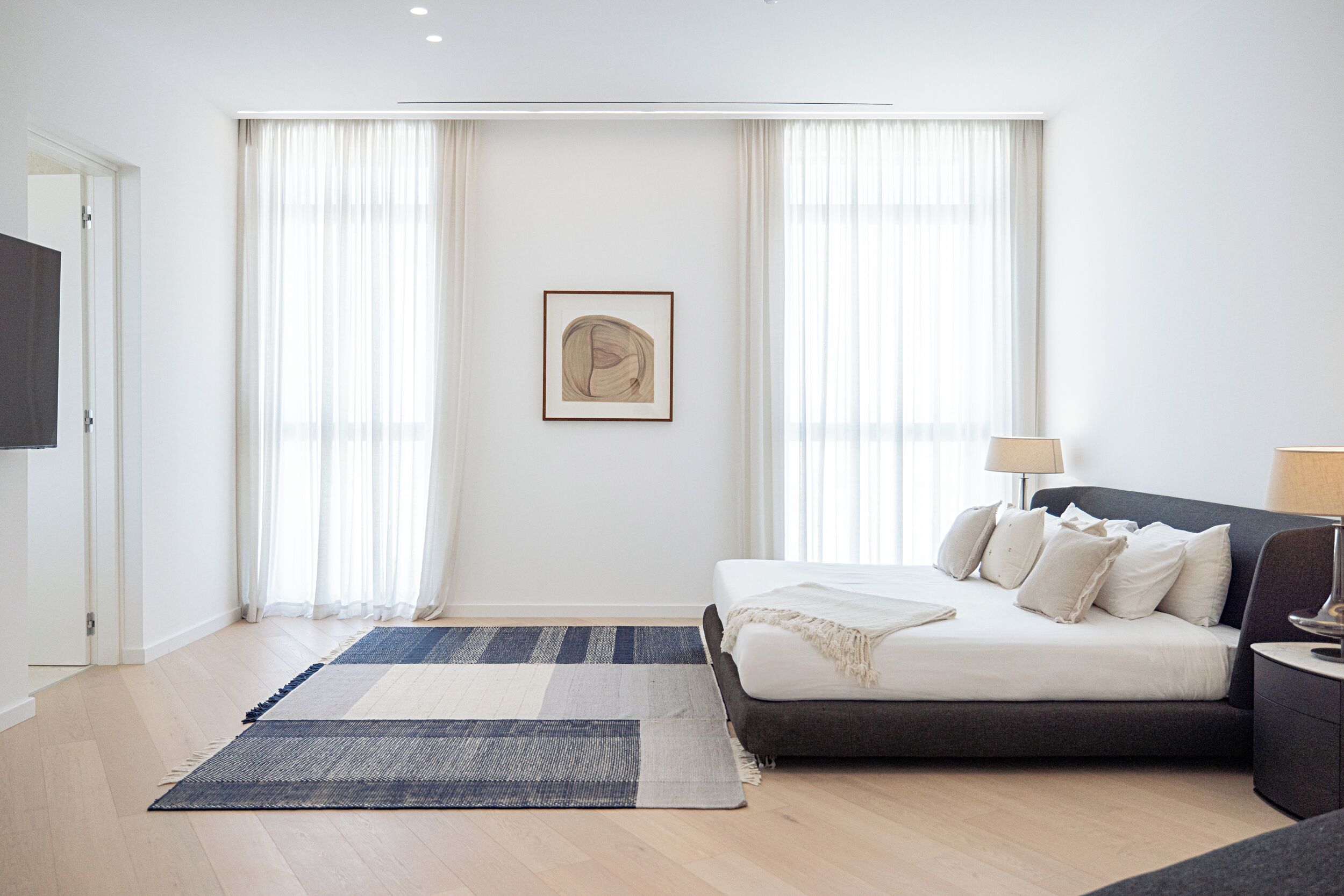 Un dormitorio moderno con cama, mesitas de noche, televisor y un amplio ventanal con cortinas.