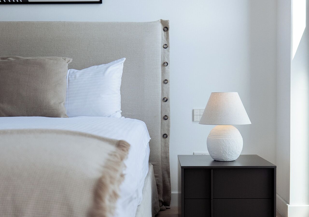 A neatly made bed with a grey headboard, a white pillow, and a beige throw blanket. There is a white textured lamp on a black bedside table beside the bed.