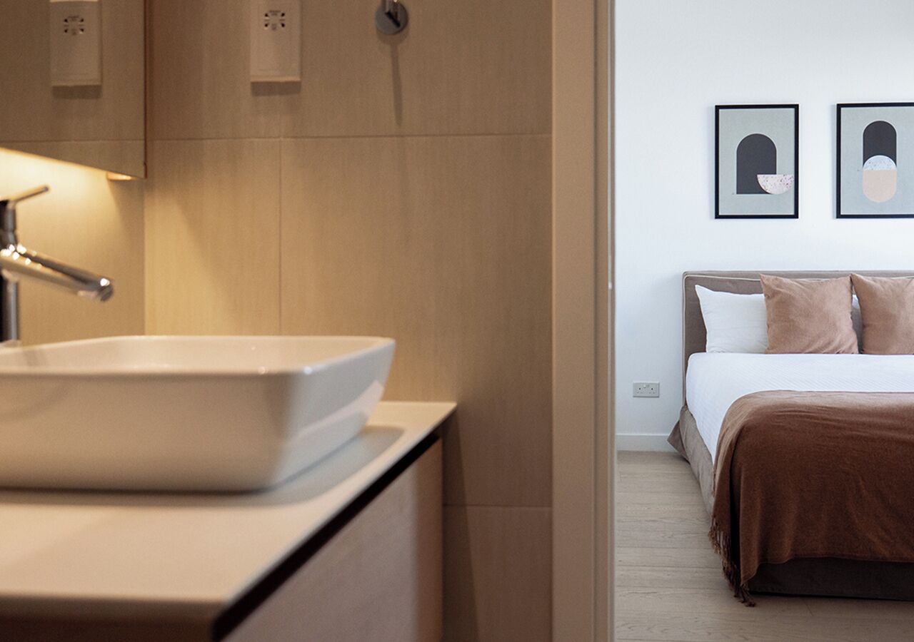 Une salle de bain moderne avec un lavabo blanc et une table de chevet. La pièce contient un lit recouvert d’une couverture marron et deux tableaux encadrés accrochés au mur.