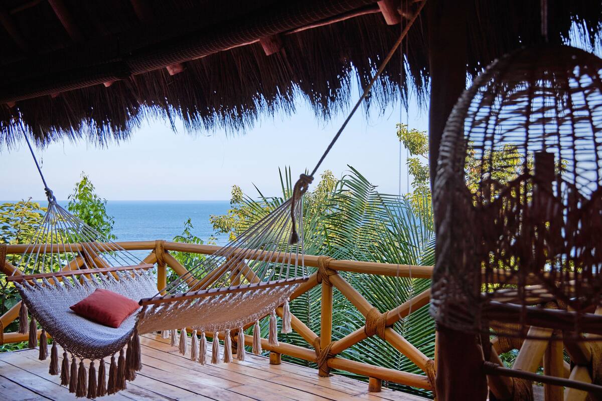 A hammock chair with a red cushion on a wooden deck overlooking the sea.
