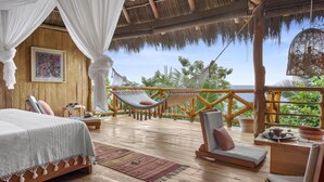 A wooden deck with a bed, two hammocks, and a seating area overlooking a lush green landscape and the sea.