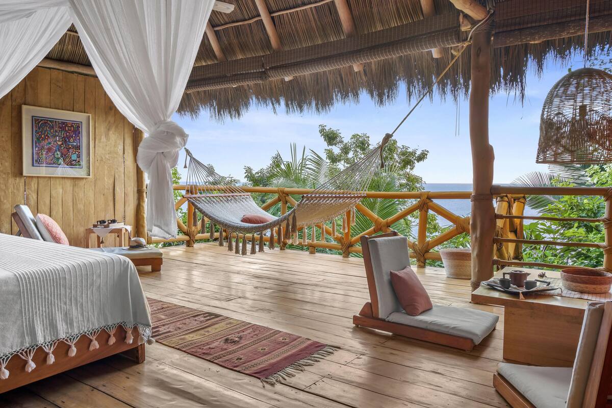 A wooden deck with a bed, two hammocks, and a seating area overlooking a lush green landscape and the sea.
