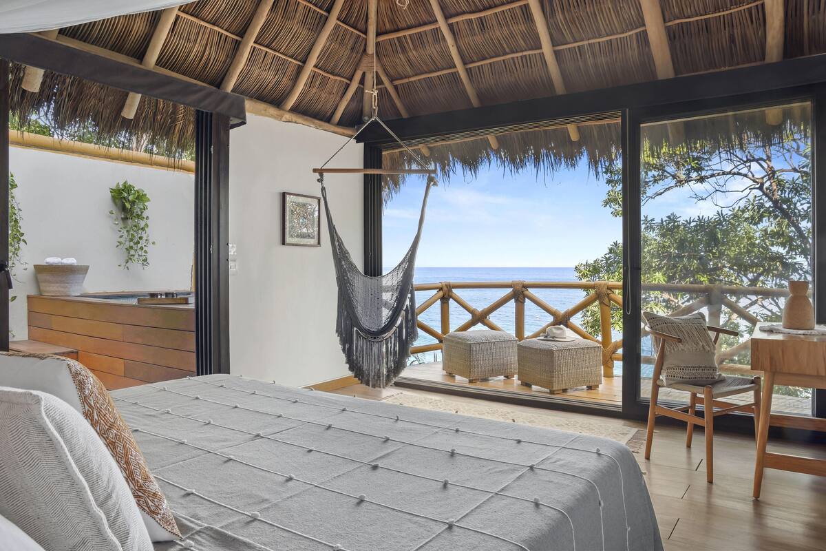 A bedroom with a hammock, a bed, a wooden table, and a view of the sea.