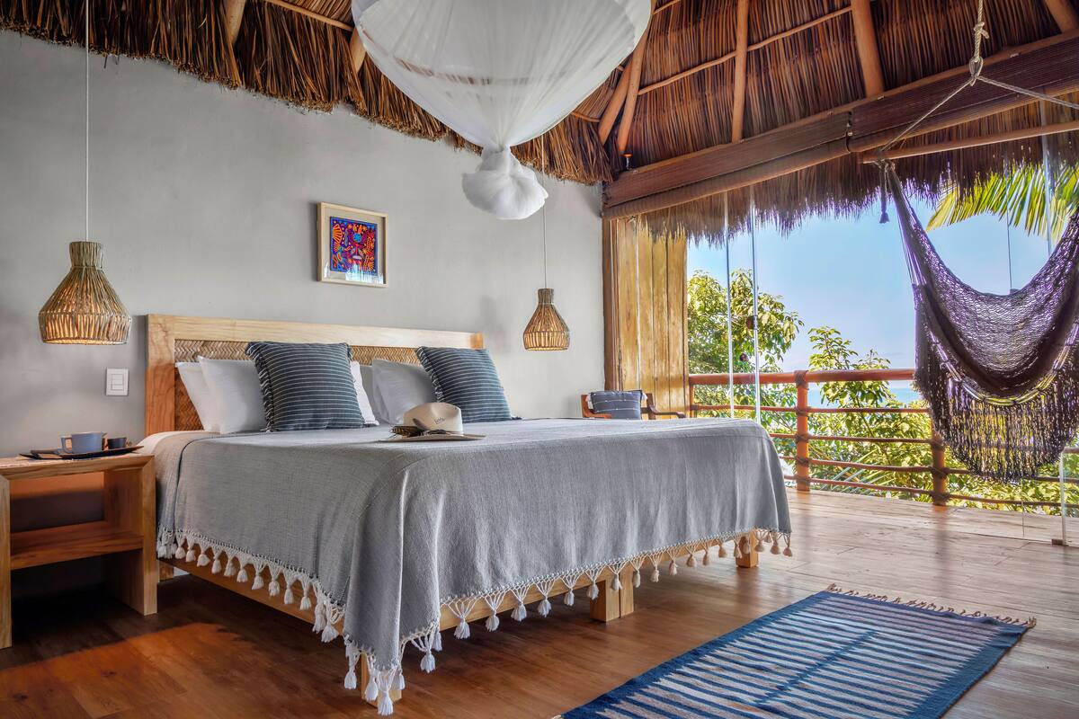 A bedroom with a bed, bedside tables, a hanging chair, and a view of the outdoors.