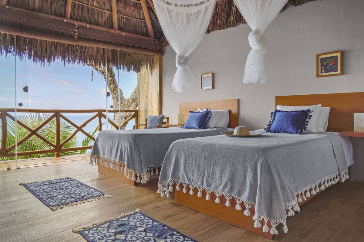 A room with two beds, a wooden floor, and a balcony with a view of palm trees.