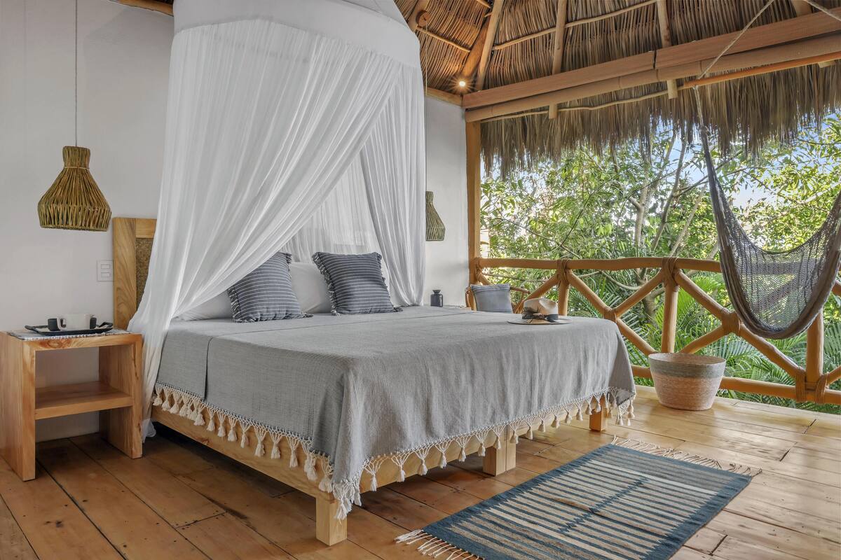 A bed with a canopy, wooden flooring, a nightstand, a hanging light, and a view of greenery outside.