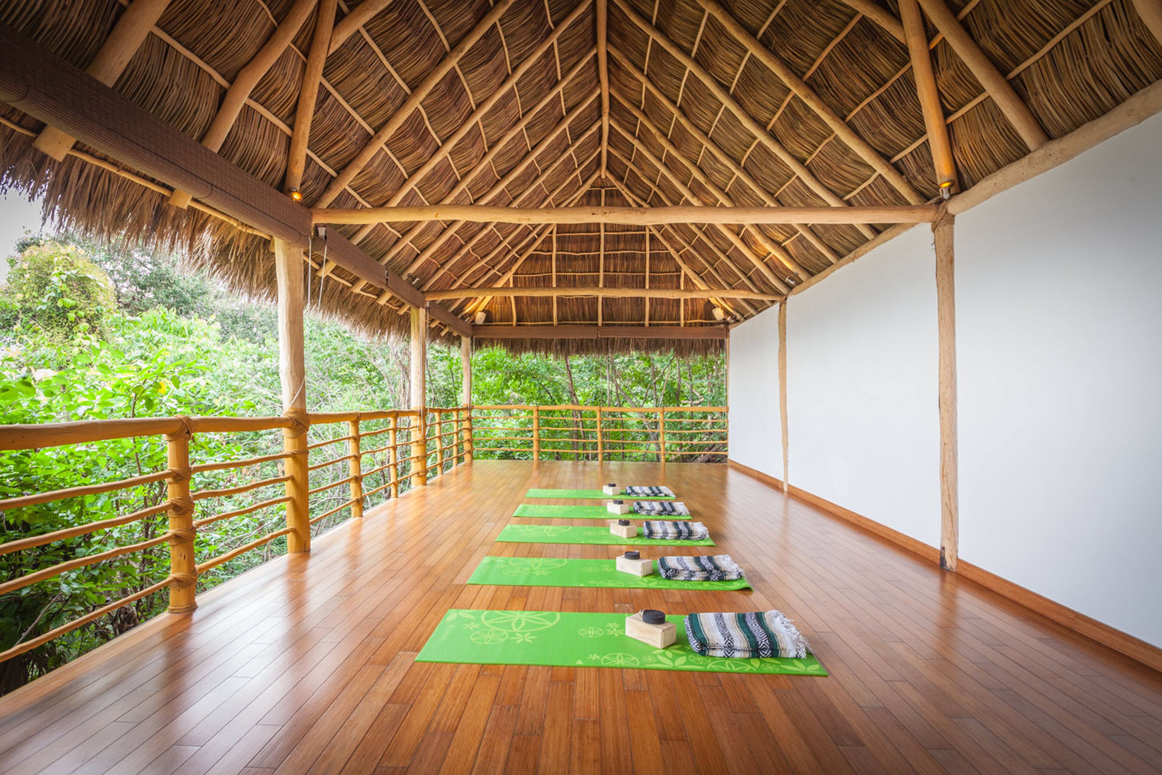 Una terraza de madera con techo de paja, tapetes de yoga verdes y bloques de madera dispuestos para la práctica.