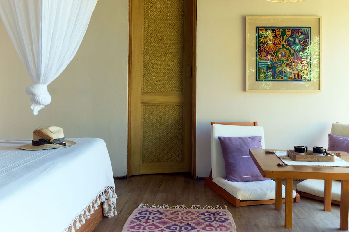 A room with a bed, a chair, a table with cups, a hanging light fixture, and a door with a woven screen.