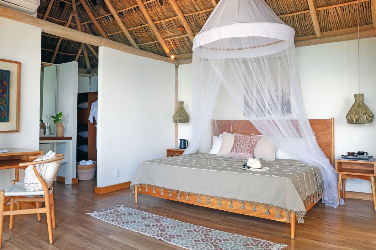 A room with a bed under a canopy, a desk with a chair, a small table with cups, and a woven lamp.
