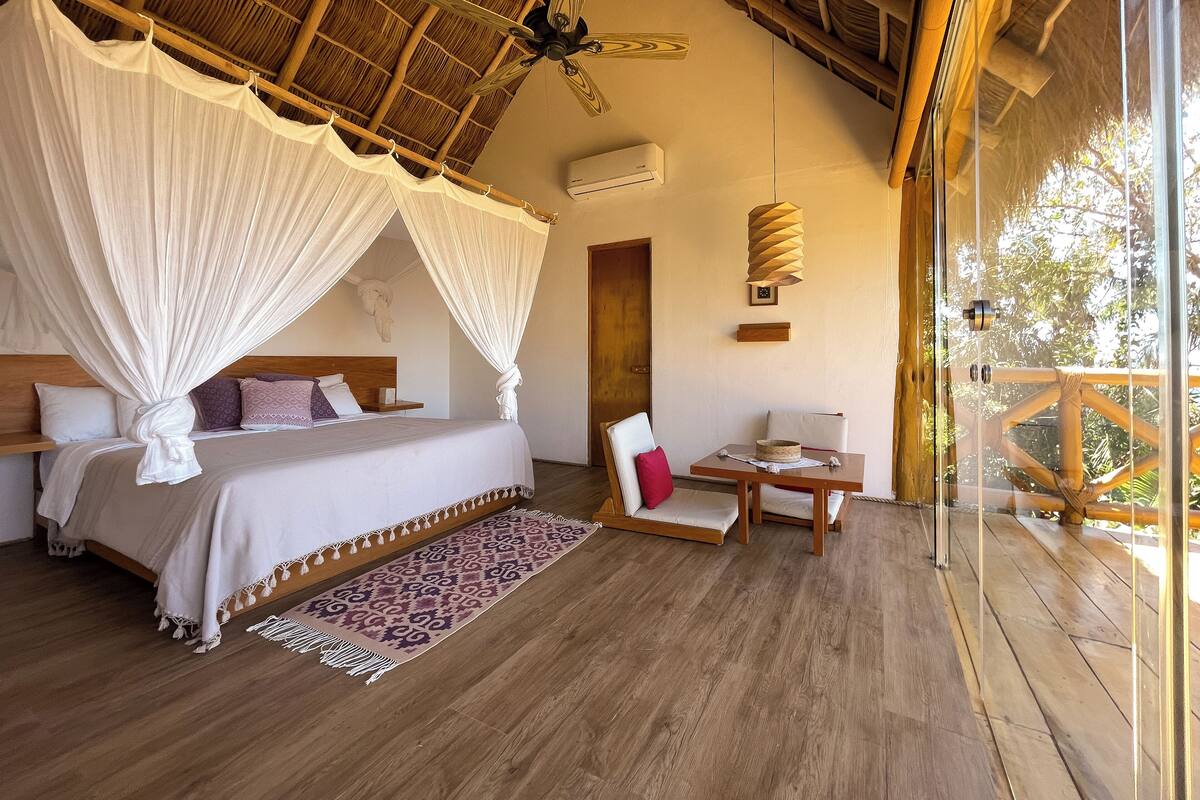 A bedroom with a canopy bed, a wooden ceiling, a ceiling fan, a table with chairs, and a view of the outdoors.