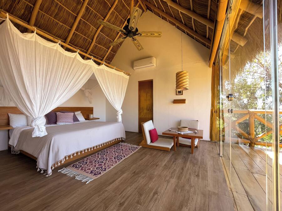 Un dormitorio con cama con dosel, techo de madera, ventilador de techo, una mesa con sillas y vistas al exterior.