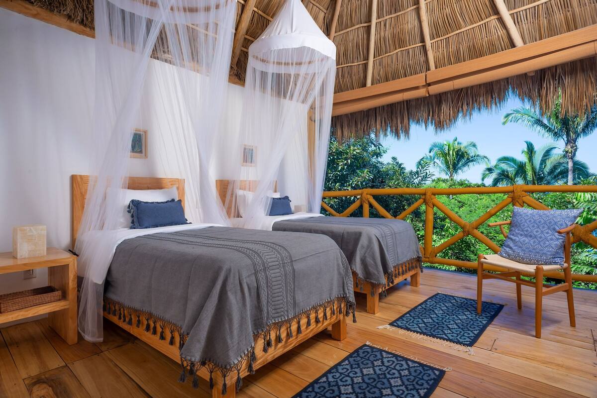 A room with two beds, a wooden floor, a thatched roof, and a view of palm trees.
