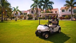 Un carrito de golf sobre un césped cuidado, con palmeras y un edificio al fondo.