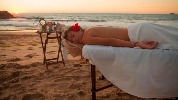 Una persona recostada sobre una camilla de masaje en la playa, con un atardecer de fondo.