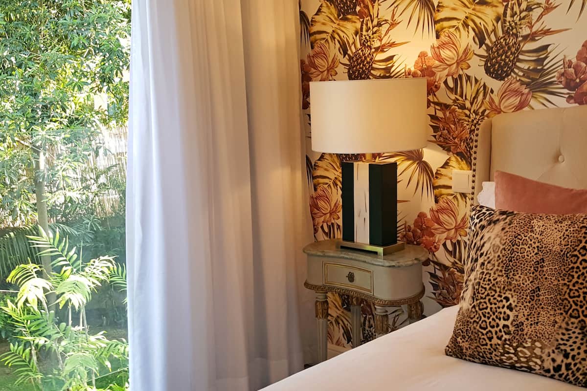 Ein Bett mit Zierkissen mit Leopardenmuster, ein Nachttisch mit Lampe und blumiger Wandtapete.