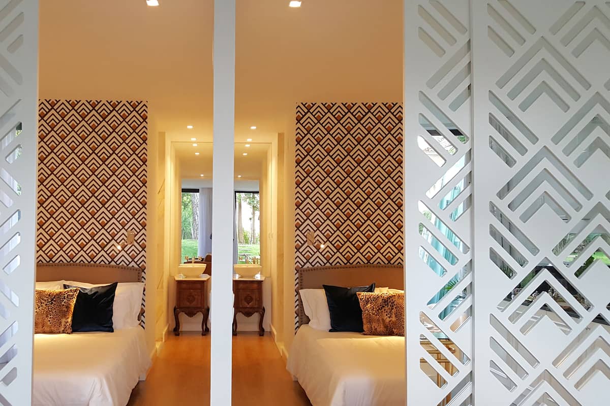 Ein Zimmer mit Spiegeln und einer Wand mit geometrischem Muster, ein Bett mit weißer Bettwäsche, ein Nachttisch und ein Fenster mit Blick auf Grün.