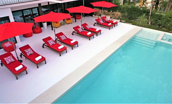 A poolside area with red lounge chairs and umbrellas, a clear blue pool, and a view of greenery and buildings in the background.