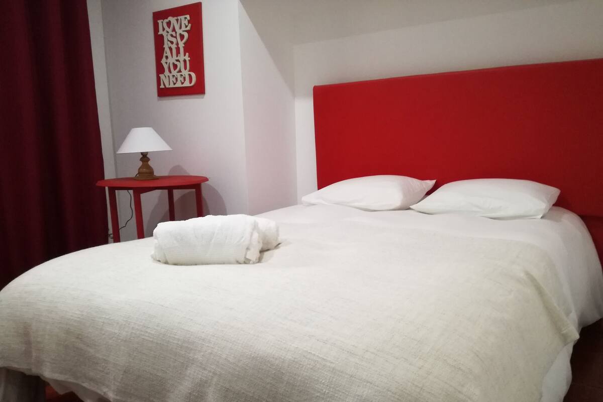 Una cama bien hecha con sábanas blancas, un cabecero rojo y una mesita de noche con lámpara.