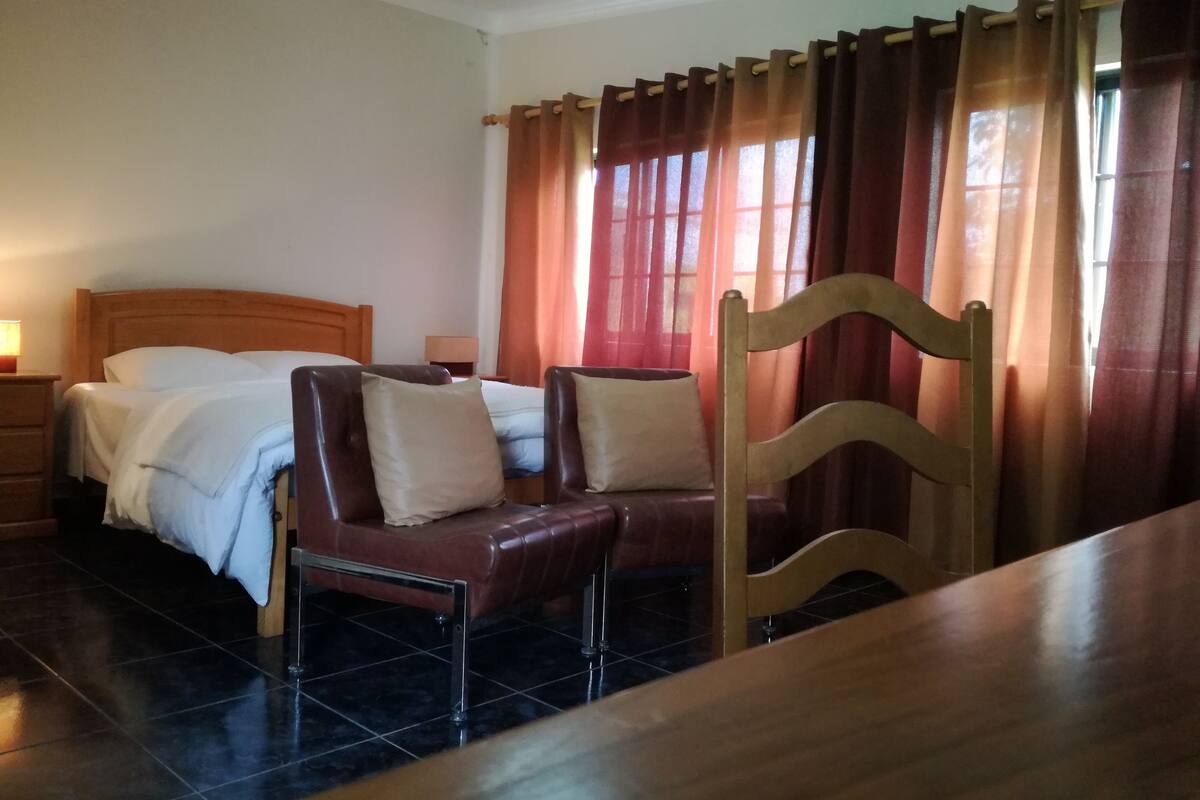 Habitación de hotel con una cama de madera, dos mesitas de noche, una silla de madera, una mesa de comedor de madera y una ventana con cortinas.