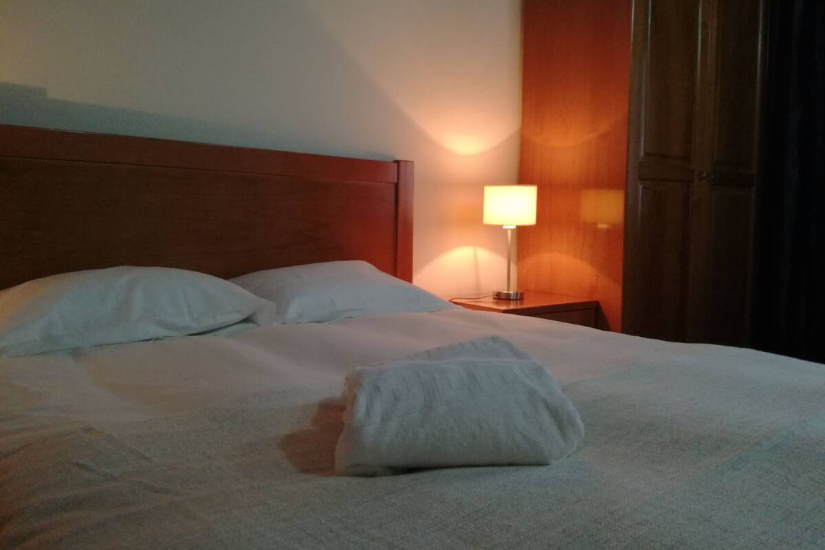 Una cama bien hecha con sábanas blancas, un cabecero de madera y una lámpara de noche.