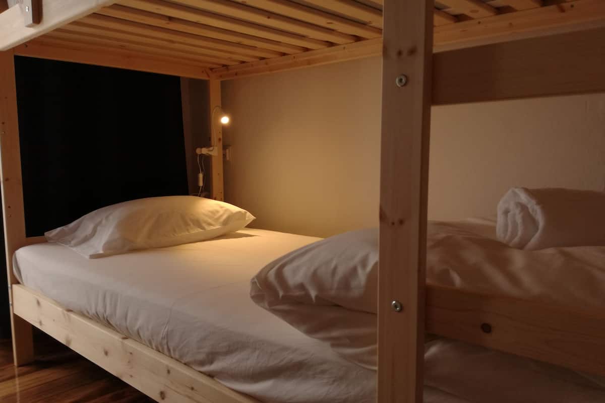 Una cama litera con ropa blanca, cabecera de madera y una mesita de noche con lámpara.