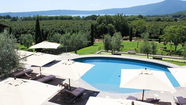 Piscina con sillas de descanso y sombrillas, rodeada de un jardín ajardinado y vistas a un lago y montañas.