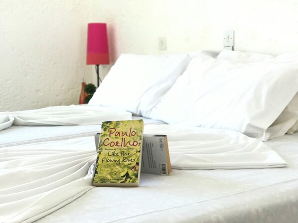 Una cama bien hecha con sábanas blancas, un libro titulado 'Como el río fluye' de Paulo Coelho y una lámpara rosa al fondo.