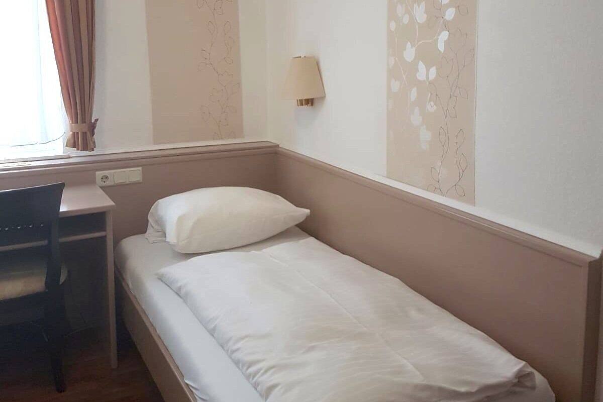 Un petit lit bien fait, avec une literie blanche, dans une pièce qui contient un bureau et une chaise en bois.