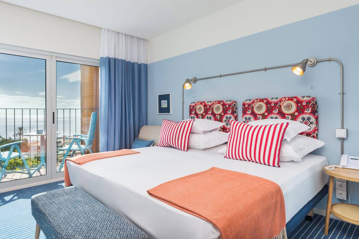 Une chambre d’hôtel avec un lit, une table de chevet avec un téléphone, une fenêtre avec des rideaux et un balcon avec des chaises.