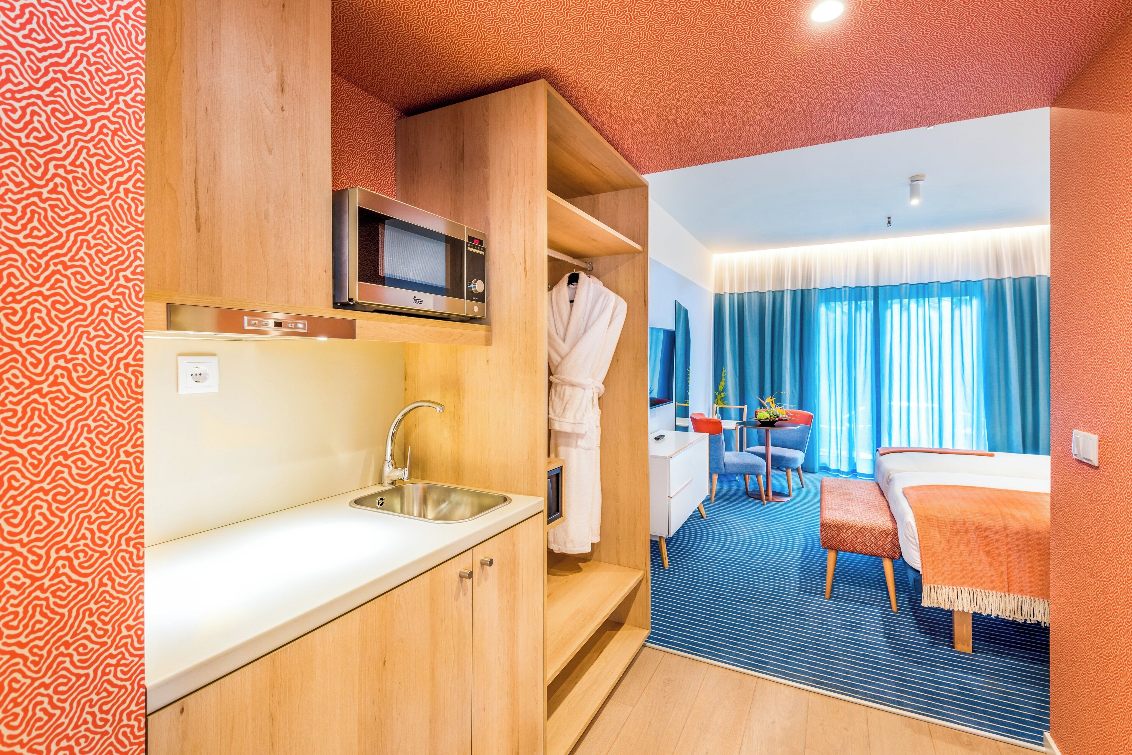 A compact hotel room with a kitchenette, a bed, a small table, and a chair.