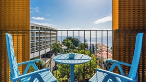 Un balcón con dos sillas azules y una mesita, con vista a un paisaje urbano con edificios y el mar.