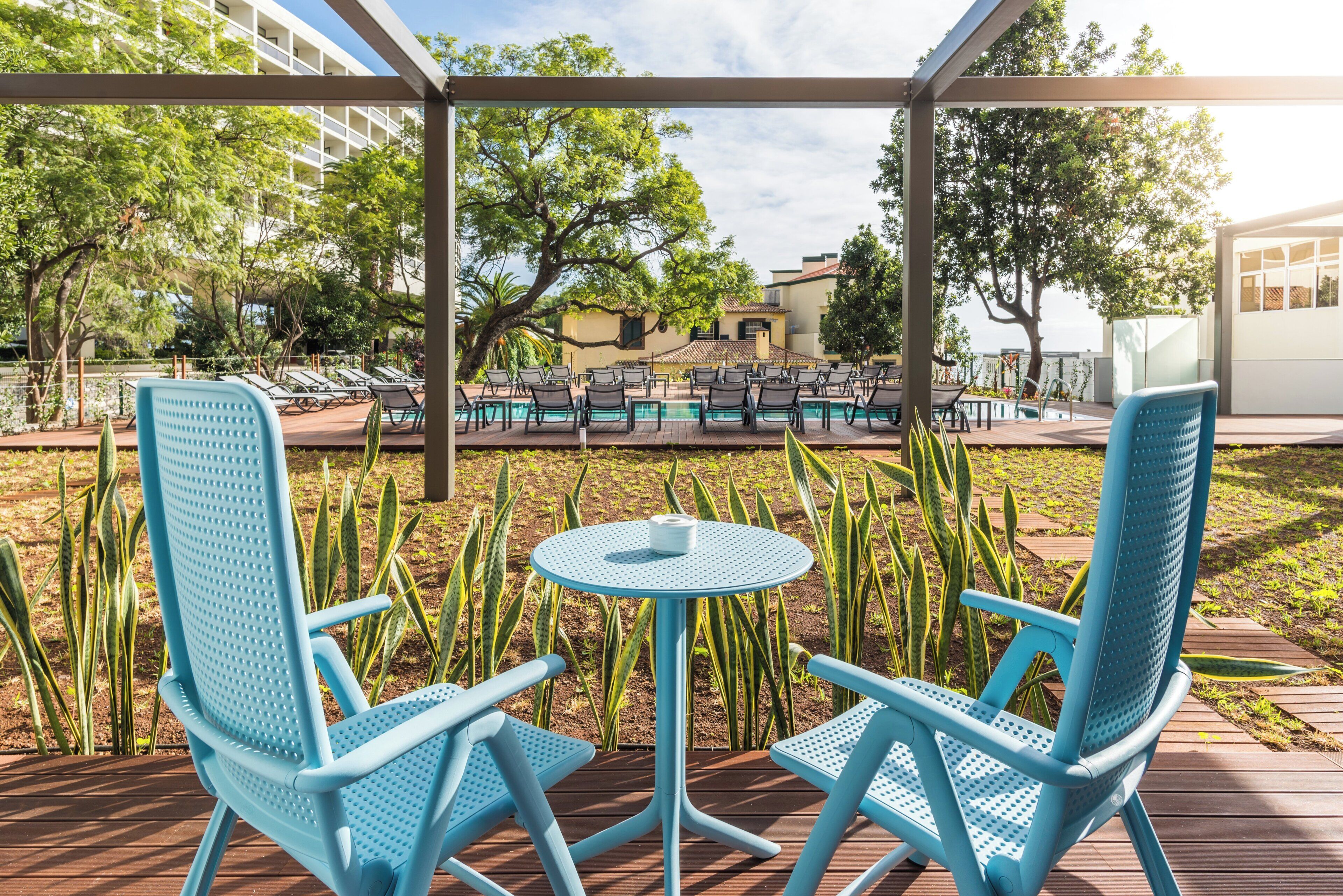 Un patio con sillas azules y una mesa redonda pequeña, una piscina al fondo y un edificio con balcones.