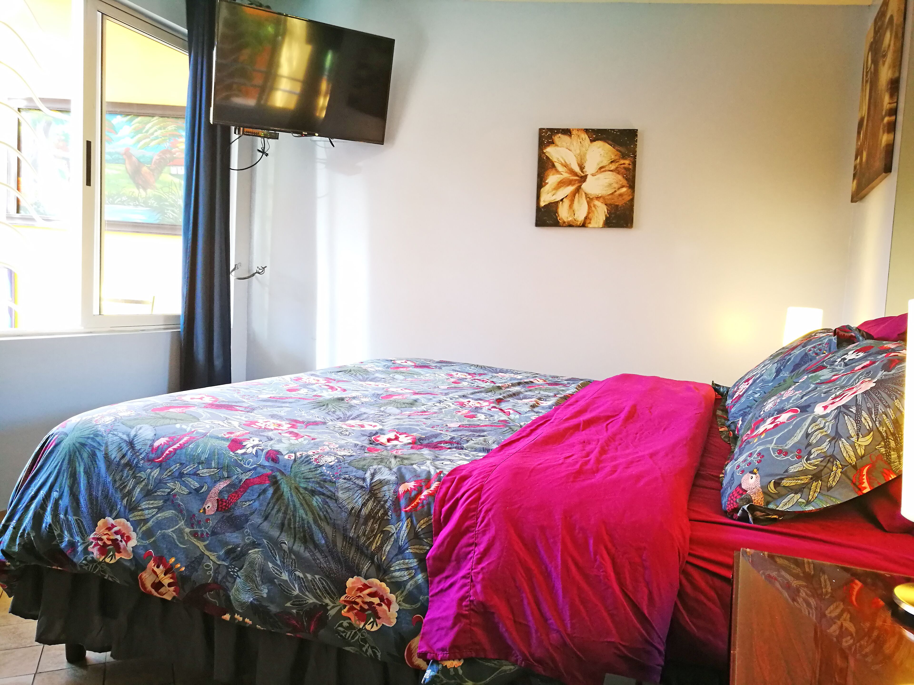 A bedroom with a bed, a TV mounted on the wall, a window with curtains, and a floral painting on the wall.