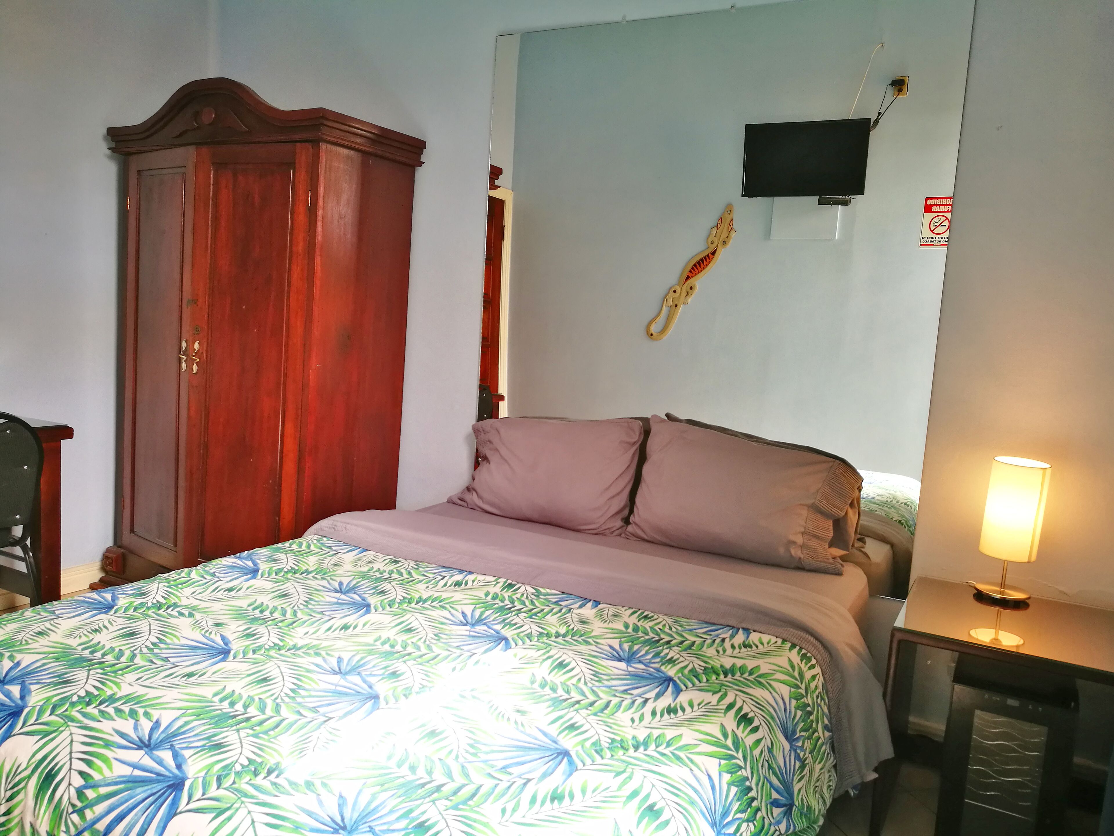A bedroom with a bed, a wooden wardrobe, a television mounted on the wall, and a nightstand with a lamp.
