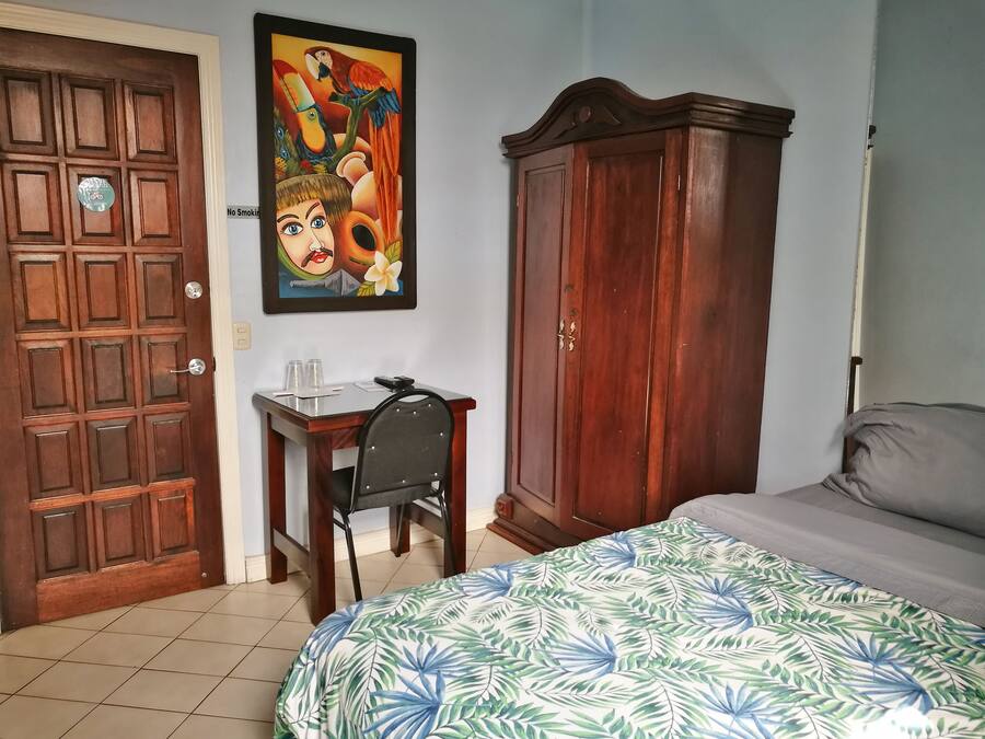 A bedroom with a bed, a wooden wardrobe, a desk with a chair, and a wall-mounted painting.