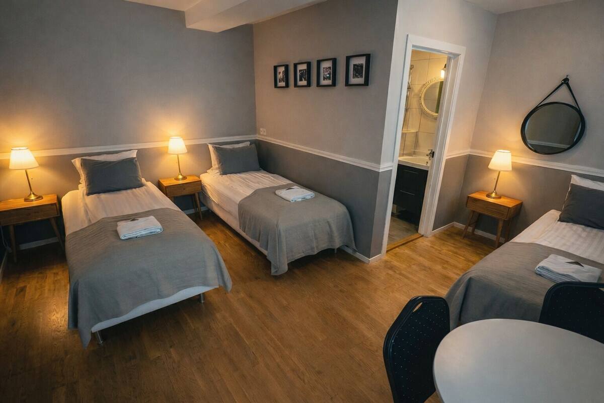 A hotel room with four single beds, each with a bedside table and lamp. There is a small table with a chair in the corner.