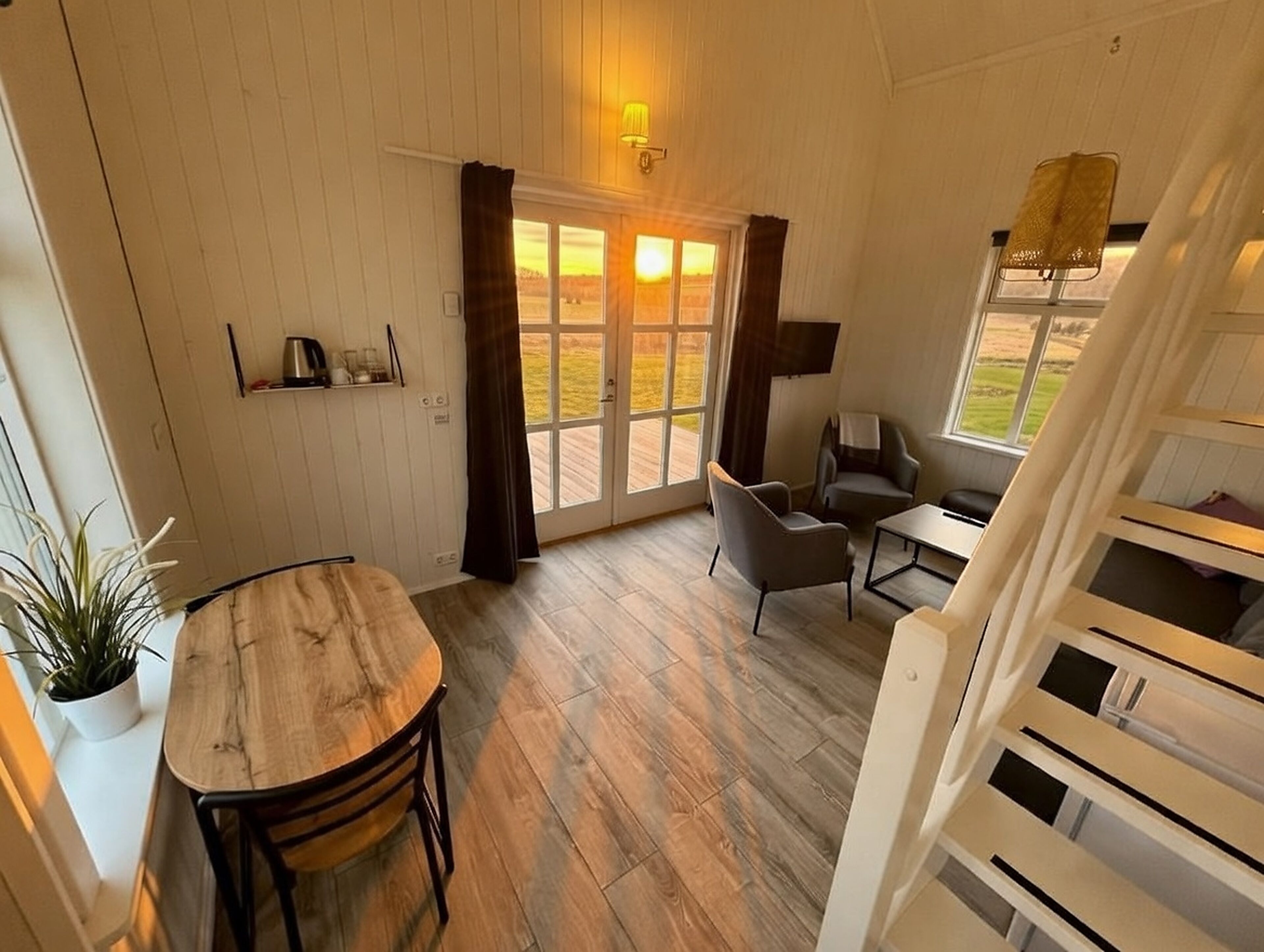 A room with a wooden dining table, two chairs, a sofa, and a small table with a lamp. There is a window with curtains and a view of the outdoors.