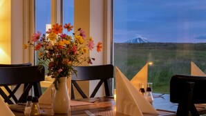 A dining table with a vase of colorful flowers, salt and pepper shakers, and a view of a mountain through large windows. - Hotel Lækur (Rangárþing ytra)