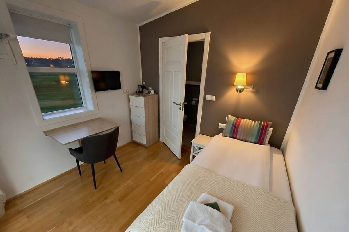A bedroom with a bed, a desk, a chair, a wardrobe, and a window with a view of the outside.
