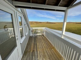 A covered wooden deck with white railings, a view of a grassy field, and a distant mountain.