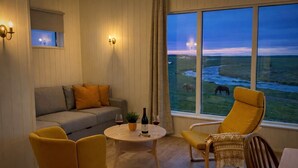 A cozy room with a sofa, two yellow armchairs, a small round table with a bottle and glass, and a view of a grassy landscape with horses. - Hotel Lækur (Rangárþing ytra)