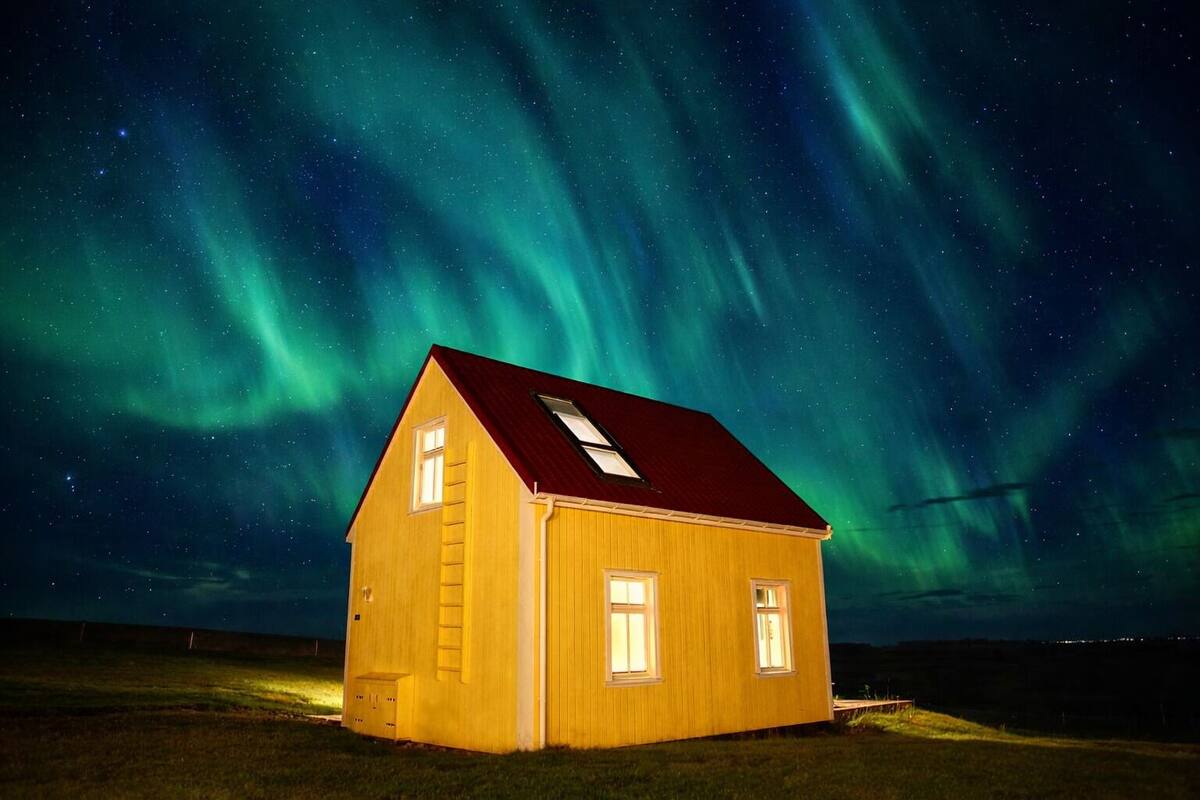 A small, single-story wooden house with a red roof and a skylight, situated under a night sky with visible aurora borealis.