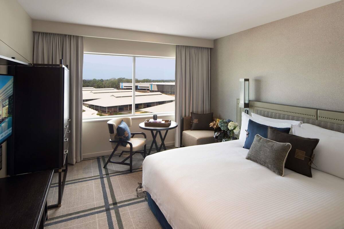 A hotel room with a bed, a desk, a chair, a TV, and a window with a view of buildings.