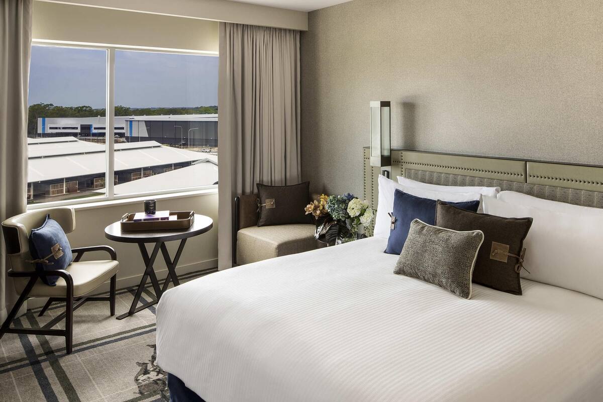 A hotel room with a large bed, a small table, a chair, and a view of buildings through the window.