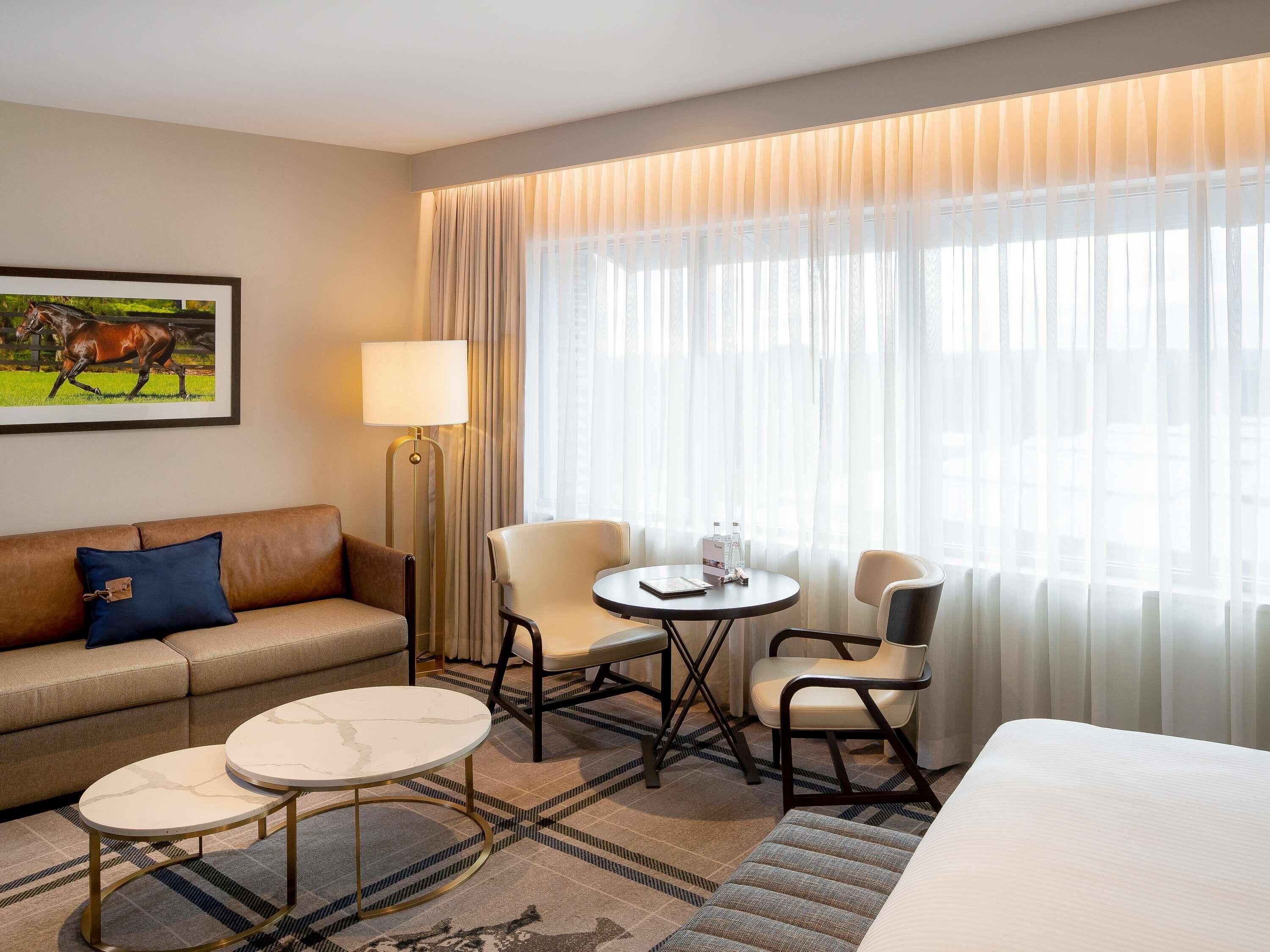 A hotel room with a sofa, two armchairs, a small table, a lamp, and a large window with sheer curtains.