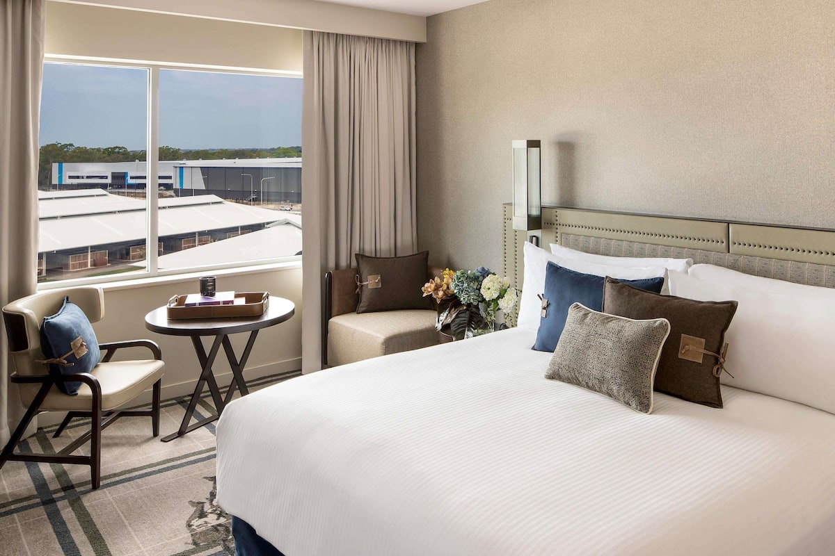 Habitación de hotel con una cama grande, una mesita pequeña, una silla y vistas a edificios a través de un ventanal.