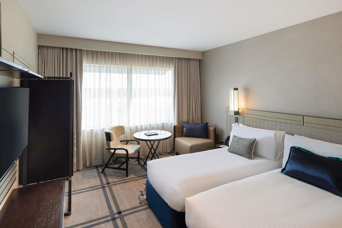 A hotel room with two beds, a desk, a chair, a television, and a window with curtains.