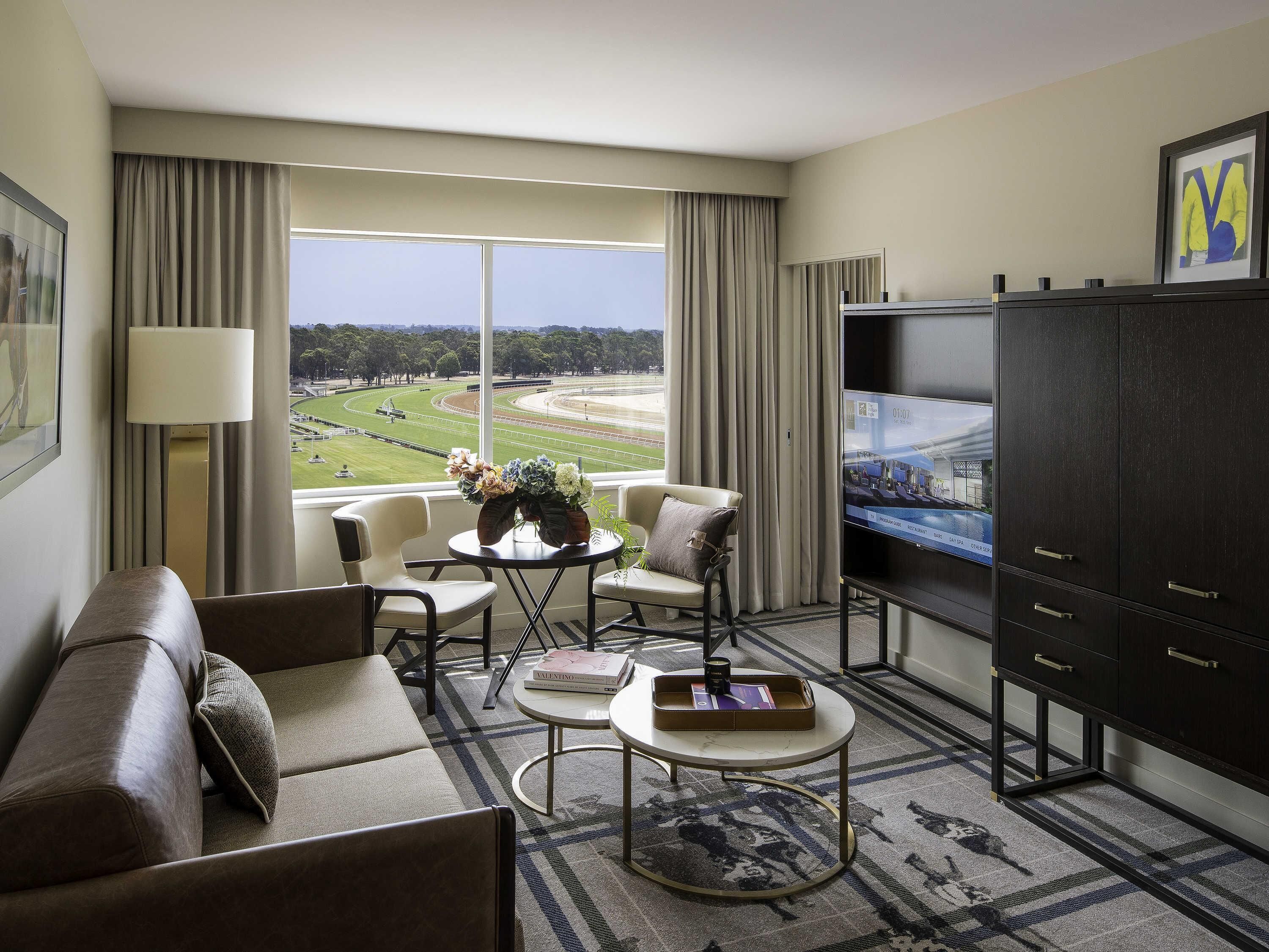 A hotel room with a large window, a sofa, a small table with a vase, a television, and a cabinet.