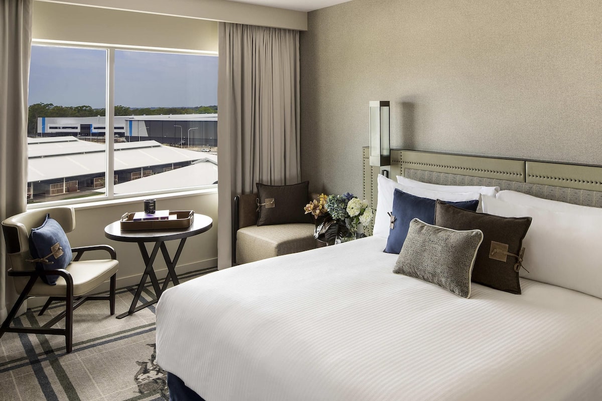 A hotel room with a large bed, a small table, a chair, and a view of buildings through the window.