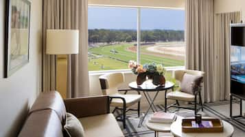 Habitación de hotel con un ventanal que ofrece vistas a una pista de carreras, un sofá, dos sillones, una mesa de centro y un televisor.
